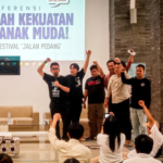 Group of young men on a stage raising their fists in celebration during a conference, with a large banner behind them reading a political youth theme in Indonesian.