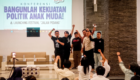 Group of young men on a stage raising their fists in celebration during a conference, with a large banner behind them reading a political youth theme in Indonesian.