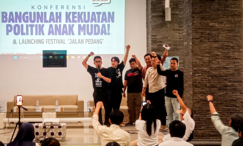 Group of young men on a stage raising their fists in celebration during a conference, with a large banner behind them reading a political youth theme in Indonesian.