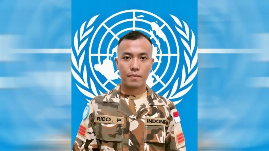 Soldier in camouflage with Indonesia flag patch standing against a United Nations emblem backdrop.