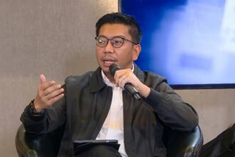 Man wearing glasses, in a white shirt and dark jacket, speaks into a handheld microphone while gesturing with his left hand during a discussion.