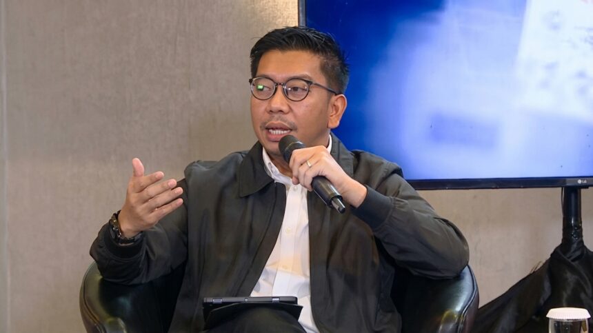 Man wearing glasses, in a white shirt and dark jacket, speaks into a handheld microphone while gesturing with his left hand during a discussion.