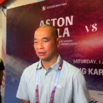 Man being interviewed at a press event, microphone in front, with a colorful backdrop advertising a match poster behind him.