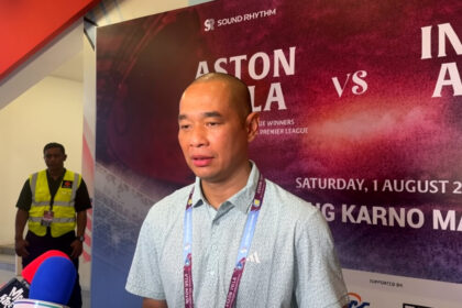 Man being interviewed at a press event, microphone in front, with a colorful backdrop advertising a match poster behind him.