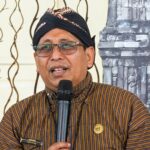Man wearing a black turban and striped brown shirt speaks into a handheld microphone at an event, wearing glasses and a round gold pin on his chest.