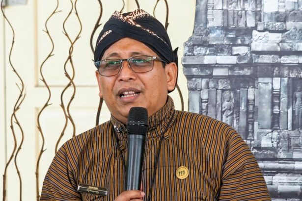 Man wearing a black turban and striped brown shirt speaks into a handheld microphone at an event, wearing glasses and a round gold pin on his chest.
