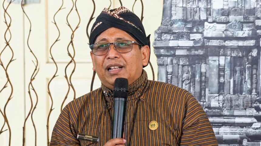 Man wearing a black turban and striped brown shirt speaks into a handheld microphone at an event, wearing glasses and a round gold pin on his chest.
