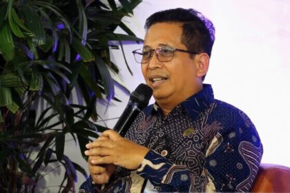 Man wearing glasses and a patterned shirt speaks into a handheld microphone, seated on a brown chair with green plants in the background.