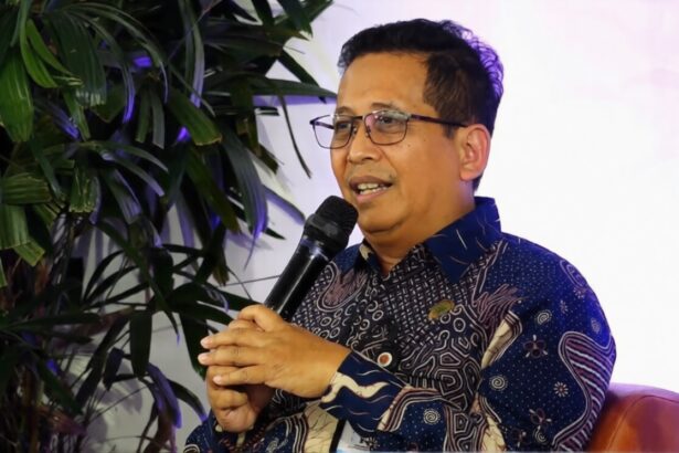 Man wearing glasses and a patterned shirt speaks into a handheld microphone, seated on a brown chair with green plants in the background.