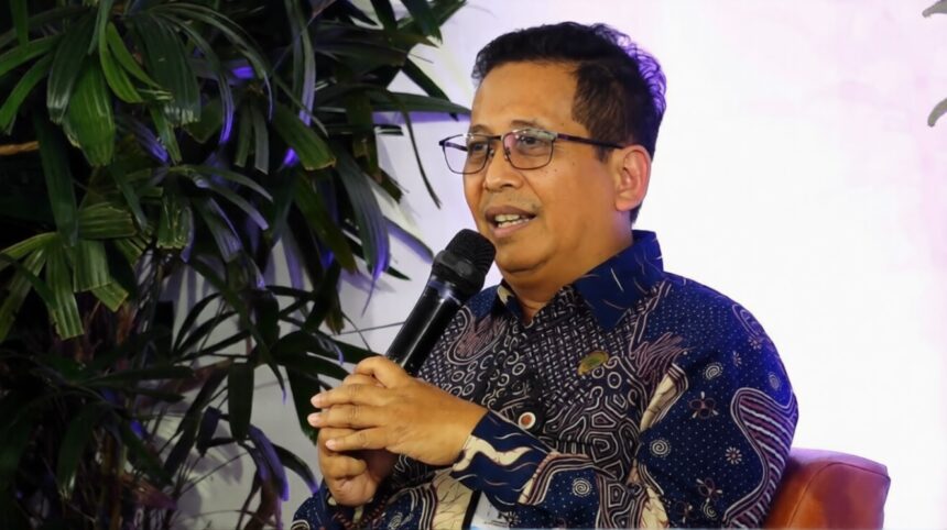 Man wearing glasses and a patterned shirt speaks into a handheld microphone, seated on a brown chair with green plants in the background.