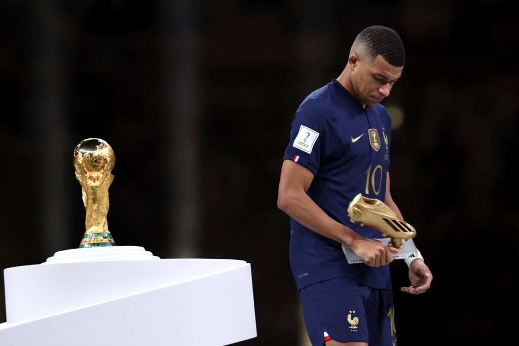 Kylian Mbappe Golden Boot GettyImages-1450107664-scaled - Mabur.co French soccer player in a dark blue France kit holds a golden boot trophy beside a pedestal with the World Cup trophy visible in the background.