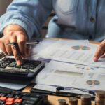 Person in a denim shirt uses a calculator and reviews financial charts on a desk