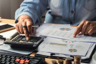 Person in a denim shirt uses a calculator and reviews financial charts on a desk