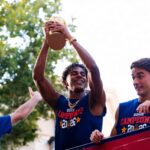 Three young men in blue jerseys celebrate on a parade float; the center player raises a trophy overhead.",