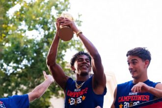 Three young men in blue jerseys celebrate on a parade float; the center player raises a trophy overhead.",