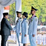 Man in a black suit shakes hands with a female officer and two male officers in light-blue dress uniforms during a formal ceremony under a tent.