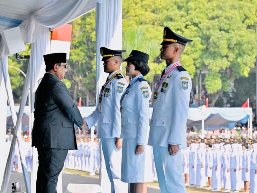 Man in a black suit shakes hands with a female officer and two male officers in light-blue dress uniforms during a formal ceremony under a tent.