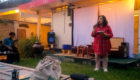 Woman in red plaid shirt speaks into a microphone on an outdoor stage at sunset event, holding papers; audience seated nearby.
