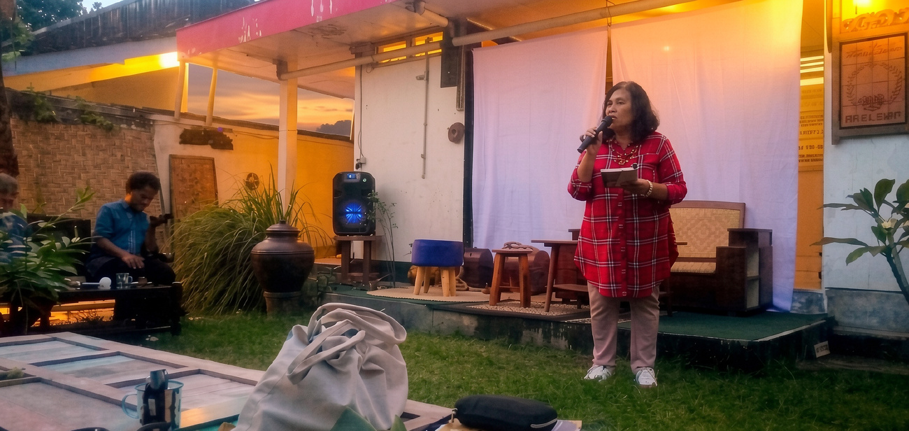 Woman in red plaid shirt speaks into a microphone on an outdoor stage at sunset event, holding papers; audience seated nearby.