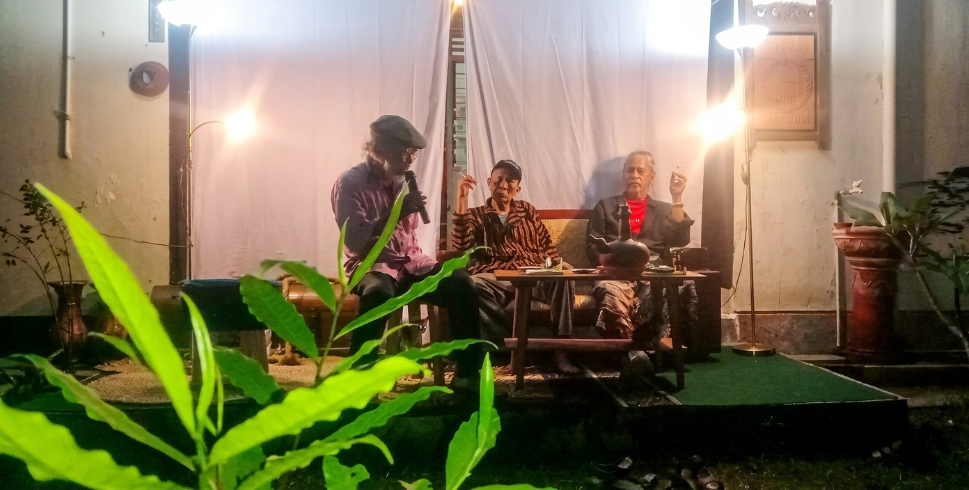 Three older men sit on a small stage with a white curtain backdrop; one speaks into a microphone while the others hold drinks, at night with warm lamps.