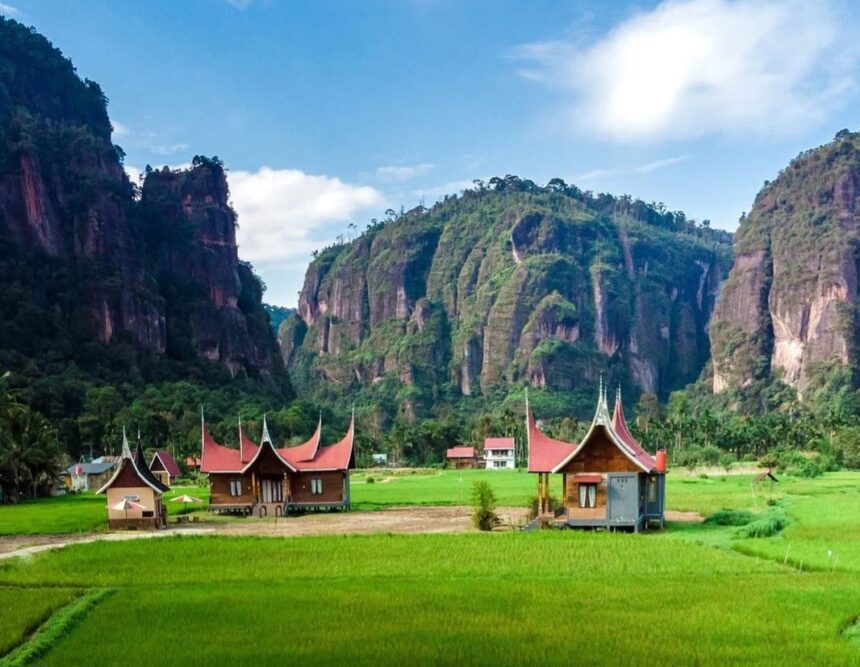 Liat deh Sob pesona sawah yang hijau dan tebing yang menjulang di LembahHarau ini bikin mata - Mabur.co Rice fields and traditional wooden houses with curved red roofs, framed by tall karst cliffs.