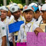 Group of students in white shirts and helmets holding colorful handmade signs at an outdoor event or parade, looking in different directions.