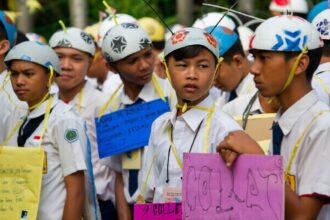 Group of students in white shirts and helmets holding colorful handmade signs at an outdoor event or parade, looking in different directions.