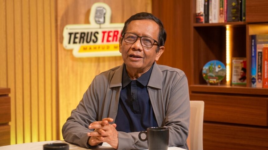 Man with glasses sits at a desk in a studio, speaking, hands clasped in front of him; microphone on his shirt and a wood-paneled backdrop behind him
