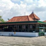 Temple-style building with red tiled roofs and white walls in a paved open area; a motorcycle parked on the right side.