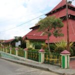 Street view of a town with a large red-roofed building behind a green and yellow fence, plants and power lines nearby.