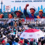 Large crowd at a labor rally facing a stage with speakers, banners reading 'Buruh Sejahtera, Indonesia Kuat' and Indonesian flags visible on stage