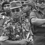 Group of soldiers in uniform, central figure wearing camouflage with sunglasses and holding a baton in front of the group.