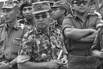 Group of soldiers in uniform, central figure wearing camouflage with sunglasses and holding a baton in front of the group.