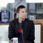 Man in a black jacket sits at a table with a microphone in front, mid-conversation during an interview in a modern studio.