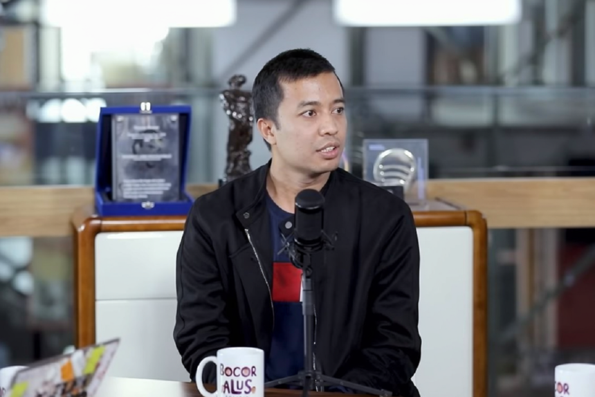 Man in a black jacket sits at a table with a microphone in front, mid-conversation during an interview in a modern studio.