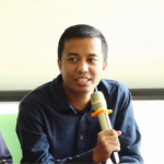 Man speaking into a handheld microphone during a panel discussion, with another man looking down beside him in a bright room