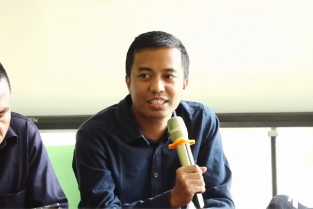Man speaking into a handheld microphone during a panel discussion, with another man looking down beside him in a bright room