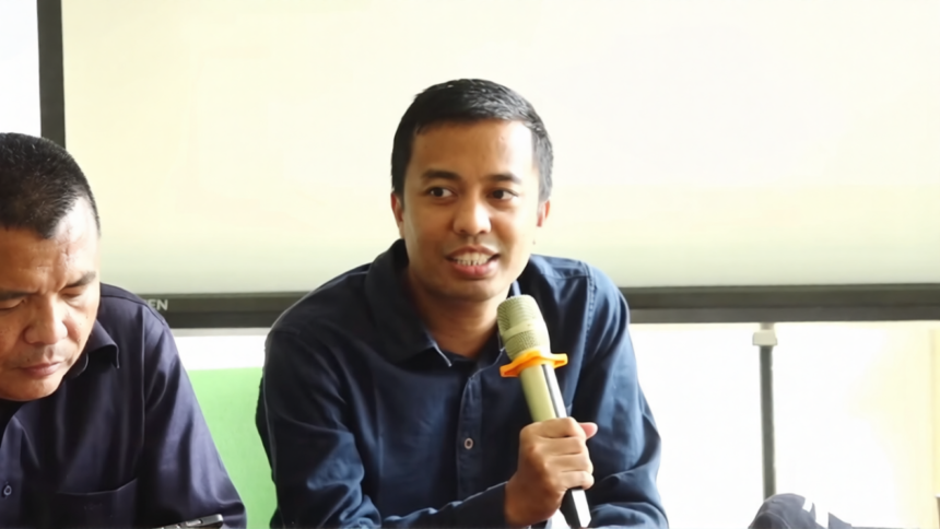 Man speaking into a handheld microphone during a panel discussion, with another man looking down beside him in a bright room