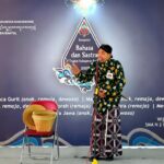 Male performer in a green batik shirt and patterned sarong speaks into a microphone on stage at a Bahasa dan Sastra event.Backdrop features logos and event text, purple gradient with decorative designs.