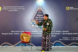 Male performer in a green batik shirt and patterned sarong speaks into a microphone on stage at a Bahasa dan Sastra event.Backdrop features logos and event text, purple gradient with decorative designs.