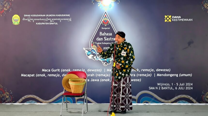 Male performer in a green batik shirt and patterned sarong speaks into a microphone on stage at a Bahasa dan Sastra event.Backdrop features logos and event text, purple gradient with decorative designs.