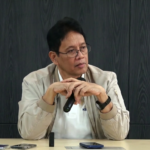 Man with glasses and a beige jacket sits at a table, holding a microphone and looking to the side as he speaks or listens.