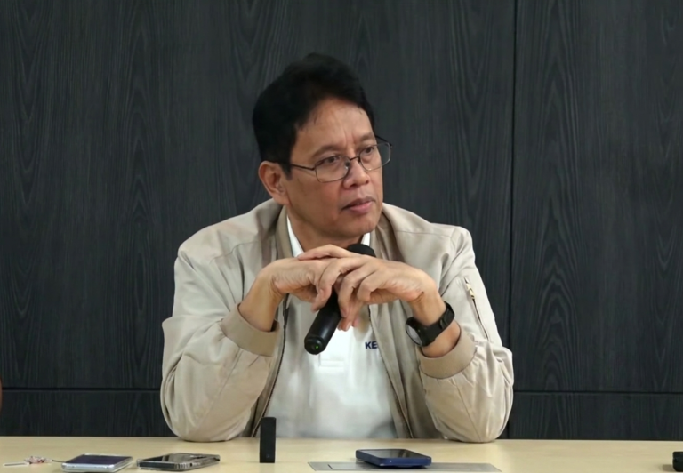 Man with glasses and a beige jacket sits at a table, holding a microphone and looking to the side as he speaks or listens.