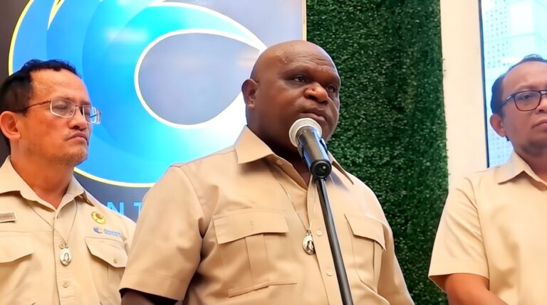 Man in tan uniform speaks into a microphone at a podium; two colleagues stand beside him against a blue logo backdrop.