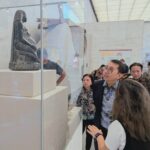 Visitors gaze at a carved seated figure displayed in a glass case at a museum, with white pedestals and a bright, marble-walled room behind them.