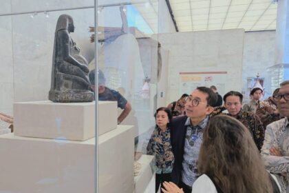 Visitors gaze at a carved seated figure displayed in a glass case at a museum, with white pedestals and a bright, marble-walled room behind them.