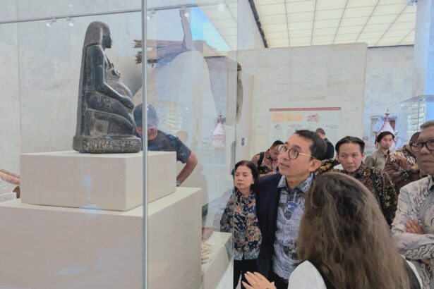 Visitors gaze at a carved seated figure displayed in a glass case at a museum, with white pedestals and a bright, marble-walled room behind them.