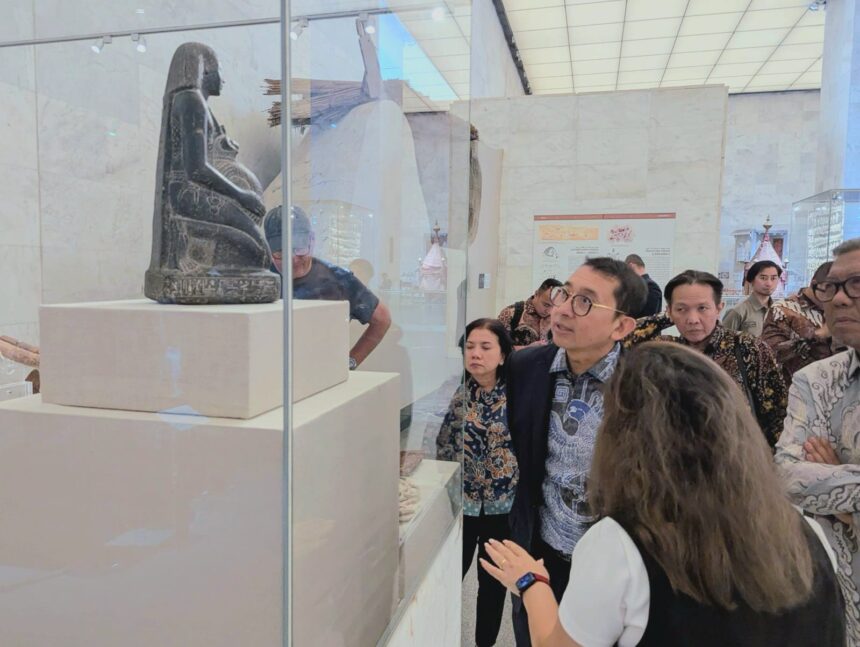 Visitors gaze at a carved seated figure displayed in a glass case at a museum, with white pedestals and a bright, marble-walled room behind them.