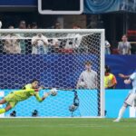 Diving goalkeeper in neon yellow makes a sprawling save near the goal as fans watch from the stands.