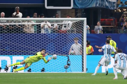 Diving goalkeeper in neon yellow makes a sprawling save near the goal as fans watch from the stands.